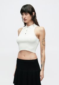 Biely rebrovaný crop top s golierovým výstrihom a čiernymi gombíkmi. Bez rukávov, zakončený jemne zakriveným lemom. Nosí sa s čiernou sukňou.