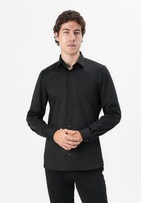 Cămașă neagră cu mâneci lungi, cu design ajustat, textură netedă și butoni negri asortați. Purtată cu pantaloni negri.