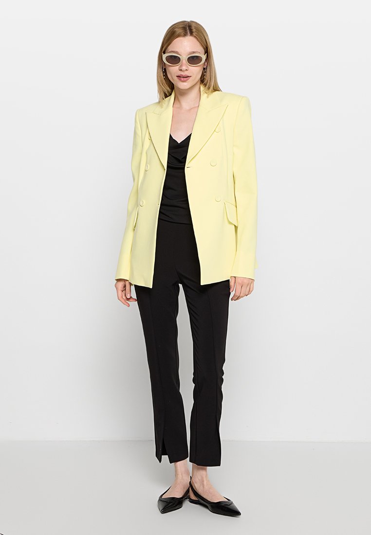 Doppelt gestreifter, hellgelber Blazer mit scharfen Revers, getragen über einem schwarzen Oberteil und einer enganliegenden schwarzen Hose, kombiniert mit spitzen schwarzen Ballerinas.
