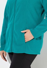Giacca in pile color turchese con zip frontale e tasche laterali, indossata da una persona con una mano in tasca e leggings neri.