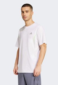 Biała sportowa koszulka krótki rękaw z okrągłym dekoltem, z szarym logo Adidas i zielonymi akcentami po bokach. Miękki, lekki materiał.
