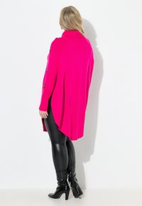 Fuksjowy, oversize sweter dzianinowy z wysokim kołnierzem, bocznymi guzikami i dłuższym tyłem, zestawiony z czarnymi legginsami z sztucznej skóry i botkami.