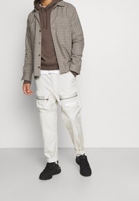 Brunrutigt skjorta, brun huvtröja under, krämfärgade cargobyxor med dragkedjor och "NIKE"-logotyp, samt svarta sneakers.