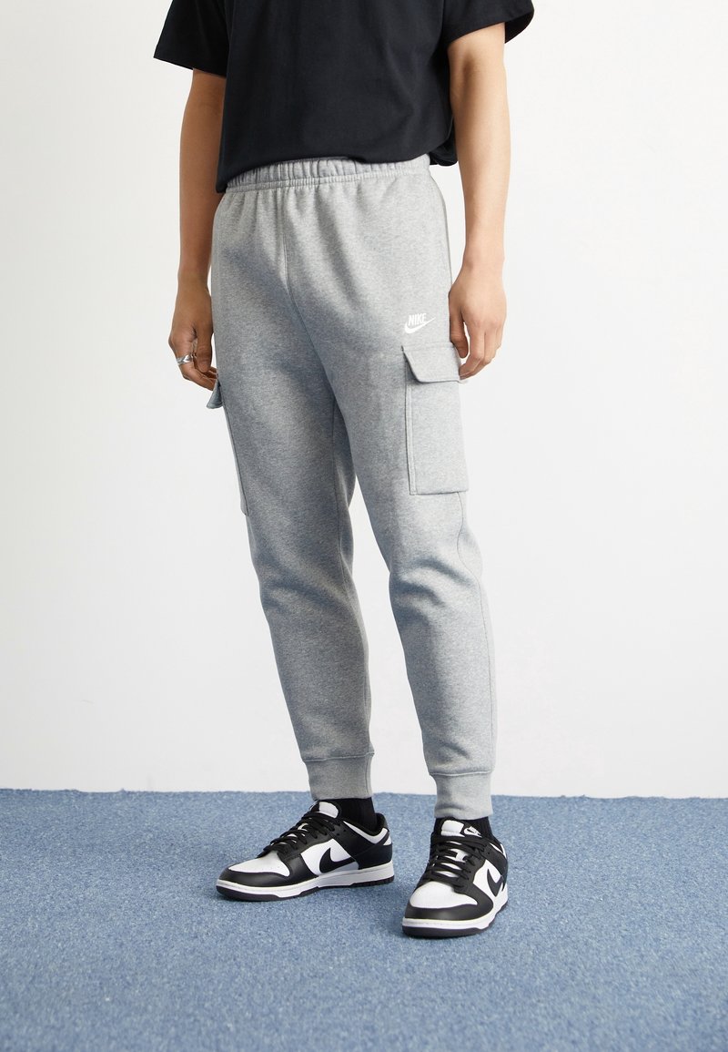 Šedé tepláky se bočními kapsami, elastickým pásem a logem Nike. V kombinaci s černobílými teniskami, na modré podlaze.