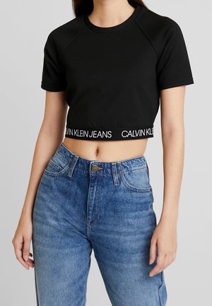 Czarny krótki t-shirt z krótkim rękawem, ozdobiony białą taśmą z logo "CALVIN KLEIN JEANS" u dołu, w połączeniu z jasnoniebieskimi spodniami z wysokim stanem.