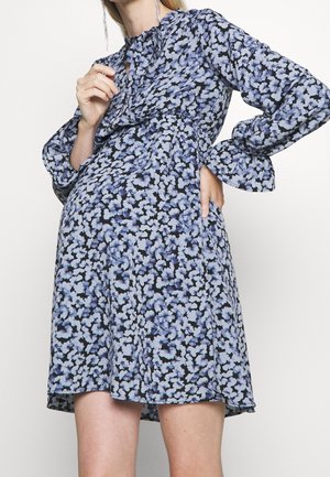 Žena v modrých a černých květovaných šatech s dlouhým rukávem a volánkovými detaily pózující s jednou rukou na boku a druhou u hrudi.