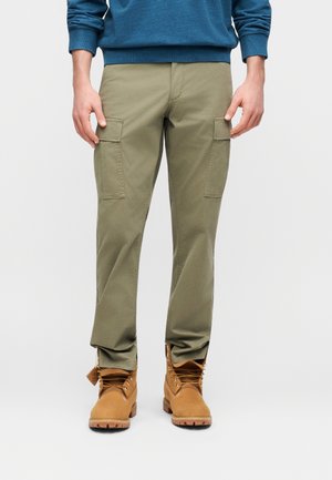 Man draagt olijfgroene cargobroek met zijzakken en beige veterlaarzen, staand tegen een effen achtergrond.