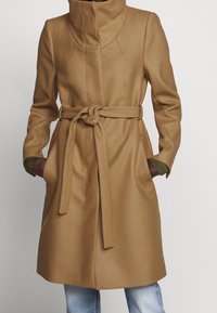 Manteau marron texturé avec un col haut, une taille ceinturée et des poches latérales. Présente une coupe ajustée et un ourlet droit.