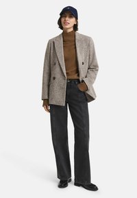 Frau, die eine marineblaue Mütze, einen braunen Rollkragenpullover, ein graues Wollblazer, schwarze weit geschnittene Jeans und schwarze Schuhe trägt, steht mit einer Hand in der Tasche.