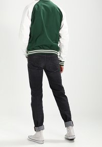 Person som bär en grön och vit varsityjacka, mörka uppvikta jeans och vita högskaftade sneakers, står med ryggen vänd mot kameran.