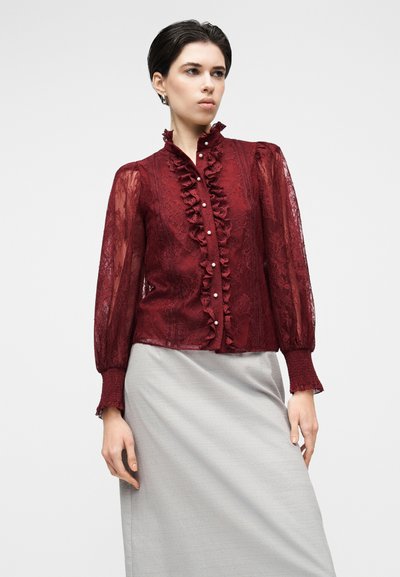 Bordová čipkovaná blúzka s volánikmi na prednej strane, vysokým golierom a priesvitnými dlhými rukávmi. Má gombíky a jemné kvetinové vzory. Kombinovaná so sivou sukňou.