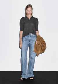 Czarna, teksturowana koszulka polo, jasnoniebieskie dżinsy z wysokim stanem i rozszerzanymi nogawkami, trzymająca brązową kurtkę z zamszu. Białe tło, minimalistyczna prezentacja.