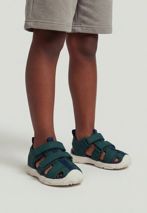 Dziecko noszące ciemnozielone i granatowe sandały zakryte z białymi podeszwami oraz beżowe szorty na jasnym tle.