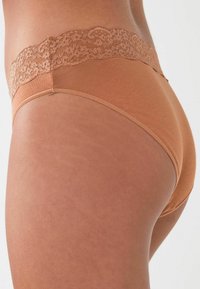 Huidkleurige katoenen ondergoed met een kanten rand rond de taille, een zachte textuur en een ontwerp dat volledige dekking biedt. Naad- en stikdetails zijn zichtbaar.