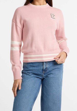 Femme portant un pull en tricot rose avec des rayures blanches aux manches et à l'ourlet, ainsi qu'un emblème en forme de lettre "G" sur la poitrine, accompagné d'un jean bleu.