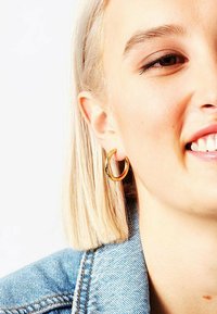 Orecchino a cerchio d'oro in esposizione, caratterizzato da un design cilindrico e liscio. Il modello ha capelli biondi lisci e indossa una giacca di jeans.