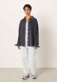 Jauna moteris stovi apsirengusi tamsiai pilką kordurovinę marškinių su sagomis, šviesiai pilką palaidinę be rankovių, šviesiai mėlynas džinsus ir pilkus bei baltus sportinius batelius.