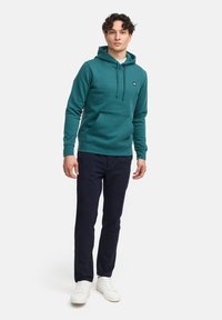 Sudadera con capucha en color verde azulado, con cordones, bolsillo canguro frontal y puños acanalados, combinada con pantalones azul marino y zapatillas blancas.