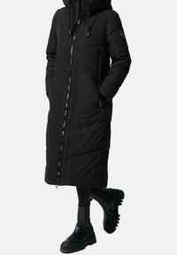 Sort lang dunjakke med lynlås foran, hætte og sidelommer. Har quiltet mønster og elastiske manchetter. Kombineret med sorte chunky støvler.