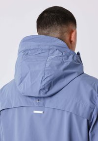 Uomo visto da dietro che indossa una giacca a cappuccio di un blu chiaro, con i capelli corti scuri e un piccolo logo metallico sulla parte superiore della schiena.
