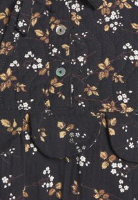 Tissu noir avec imprimé floral marron et blanc, comprenant deux petits boutons et deux poches à rabat recouvertes de tissu.