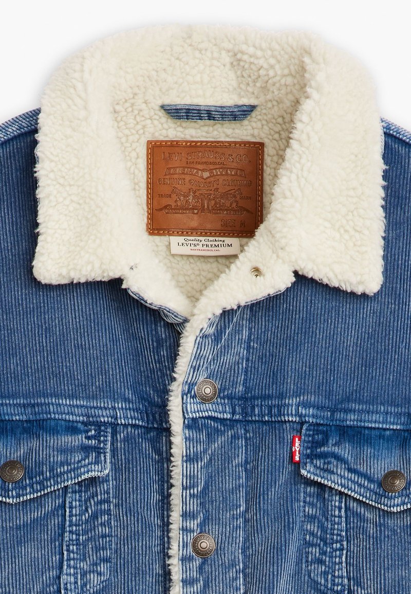 Giacca di jeans con colletto in shearling, texture in corduroy blu e etichetta in pelle. Caratterizzata da bottoni automatici in metallo e un piccolo tag rosso di Levi's.