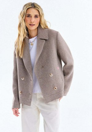 Blondynka ubrana w beżową marynarkę dwurzędową w jodełkę, białą koszulę, kremowe spodnie i warstwowe naszyjniki na jednolitym tle.