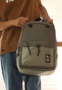 Grijze en groene rugzak met twee verstelbare banden, voorkant met zak en zwarte hardware, en logo patch. Gladde stof met subtiele textuur.