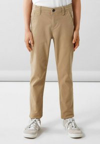 Pantaloni in misto cotone beige con vestibilità slim, dotati di tasche frontali e chiusura a un bottone. Abbinati a sneakers di colore chiaro.