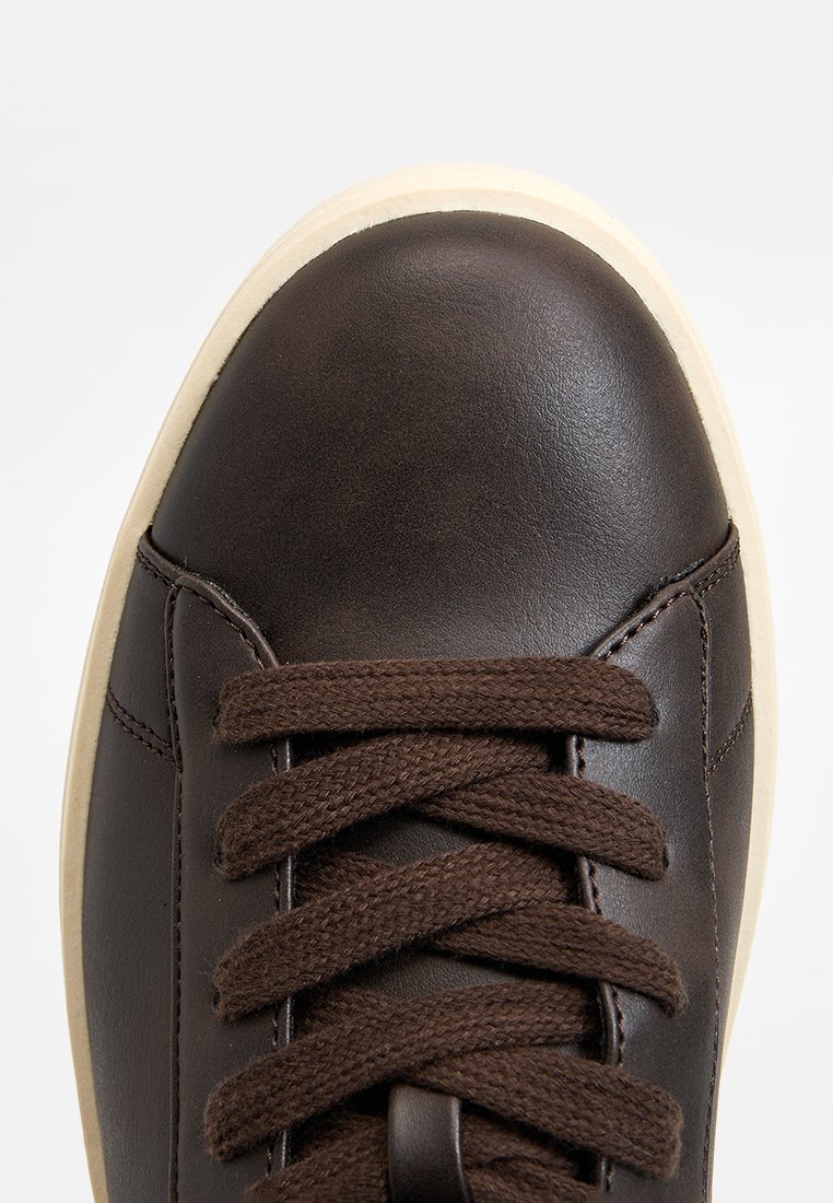 Zapatilla de cuero marrón con puntera redonda, superficie texturizada y cordones planos marrones. Suela de goma crema visible en el borde.