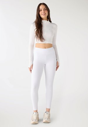 Giovane donna in piedi, che indossa un top corto bianco con cerniera, leggings bianchi a vita alta e sneakers chiari, su uno sfondo bianco semplice.