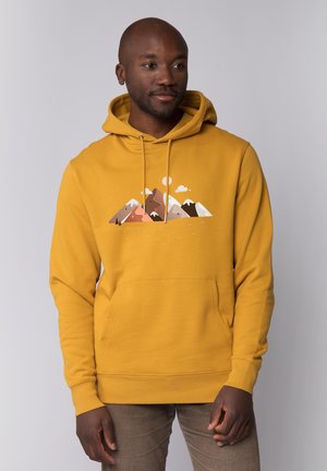 Man in mosterdgele hoodie met bergafbeelding en zon, staand met handen ontspannen langs het lichaam, licht naar rechts kijkend tegen een effen achtergrond.