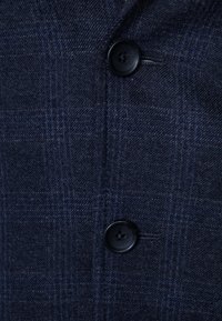 Navyblått tyg med subtilt rutmönster, som har två mörka knappar och en lätt texturerad väv. Närbild på plaggets detaljer.