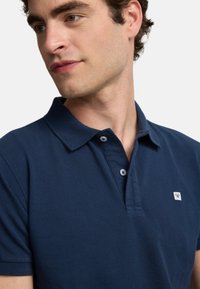 Polo de color azul marino hecho de algodón, con cuello tipo polo, tapeta de dos botones y un pequeño parche de marca en el lado izquierdo del pecho.
