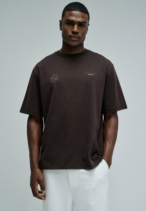 Homme portant un t-shirt marron foncé à manches courtes avec un petit texte et un motif, associé à un pantalon blanc, debout devant un fond gris uni.