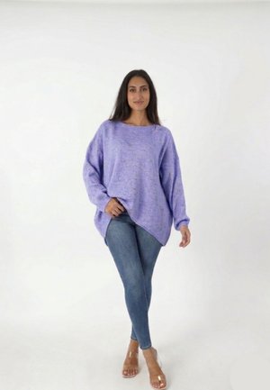 Femme debout portant un pull lavande oversize, un jean skinny bleu et des sandales à talons hauts transparentes sur fond blanc uni.