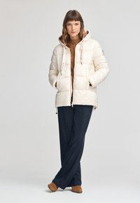 Kurtka puchowa w kremowym kolorze z kapturem, przednimi kieszeniami i akcentami logo. Teksturalne rękawy i błyszczące wykończenie. W połączeniu z ciemnymi spodniami i brązowymi butami.