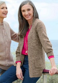Zwei lächelnde, mittelalte Frauen im Freien am Meer, die gestrickte Pullover und lässige Kleidung tragen, wobei eine auf einem Geländer sitzt.