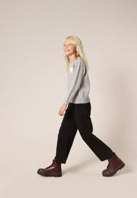 Grijze trui met een witte hartdetail, zwarte wijde broek en bordeauxrode veterschoenen met een dikke zool. Persoon wandelt op een neutrale achtergrond.