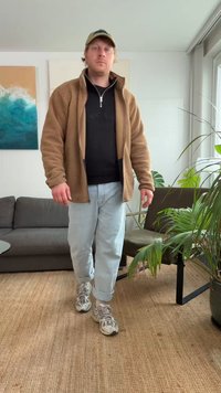 Hnedá mikina z fleecu cez čierne tričko, svetlomodré rifle a sivé tenisky. Interiérové prostredie s rastlinami a sivou pohovkou.