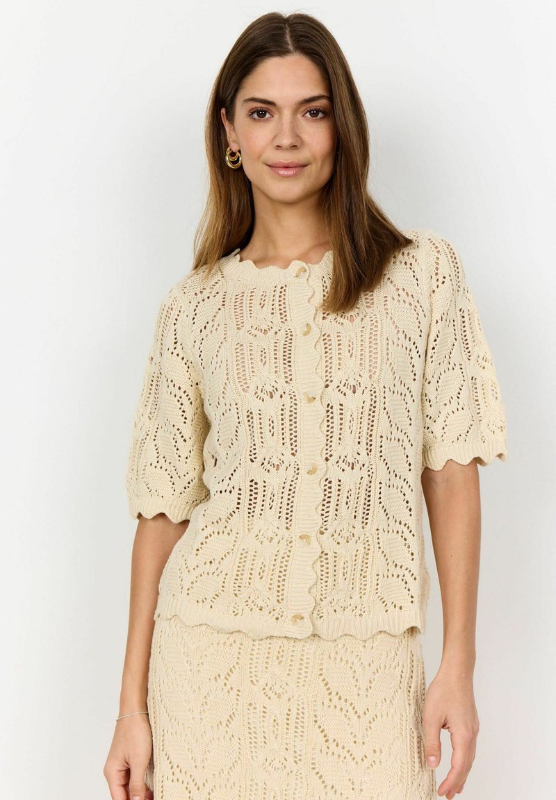 Beige gebreide top met een geschulpte halslijn en korte mouwen, voorzien van een opengewerkte patronen en knoopdetails aan de voorkant.