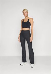 Fekete sportos crop top állítható pántokkal, fekete, testhez simuló, enyhén szárnyas szárú nadrággal párosítva. A modell fehér sportcipőt visel.
