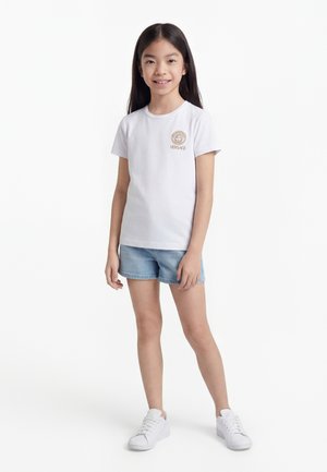 Junges Mädchen steht, trägt ein weißes Versace-Logo-T-Shirt, hellblaue Jeansshorts und weiße Sneaker, vor einem schlichten weißen Hintergrund.