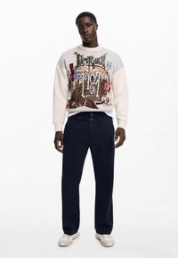 Lichtgekleurde grafische trui met ingewikkeld ontwerp, gecombineerd met donkerblauwe broek en witte sneakers, staand tegen een effen achtergrond.