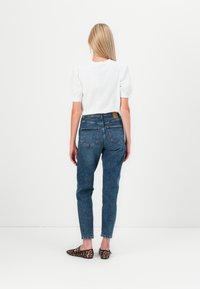 Jean en denim bleu taille haute avec une finition délavée, cinq poches et un patch en cuir marron à la taille, associé à un haut blanc à manches courtes.