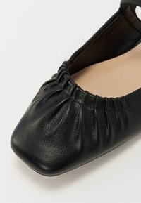 Ballerine en cuir noir avec un bout carré, mettant en avant des détails froncés le long de l'ouverture et une doublure intérieure lisse.