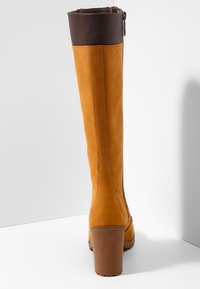 Botas de cuero hasta la rodilla en color marrón claro con un puño de cuero marrón. Cuentan con un tacón cuadrado y un cremalleras lateral, mostrando acabados suaves y texturizados.
