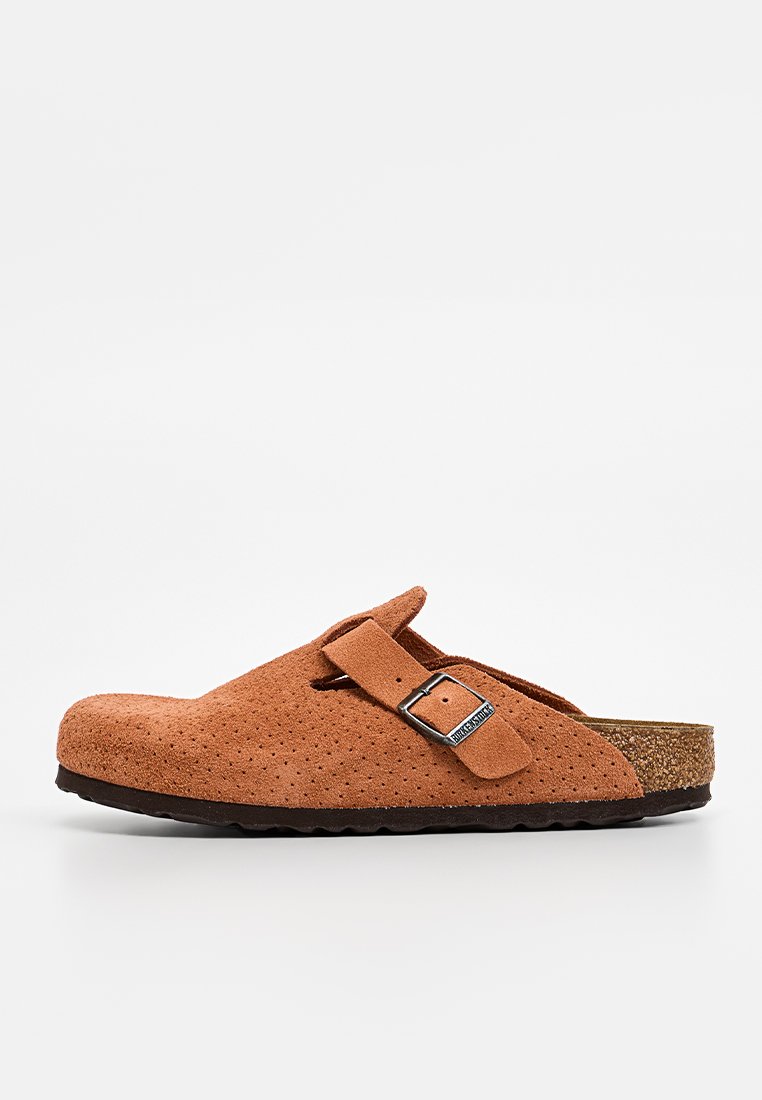 Braune Wildlederclogs mit perforiertem Obermaterial, verstellbarem Riemen und Korkfußbett. Strukturiere Sohle für Halt und Stabilität. Seitenansicht, minimalistisches Design.