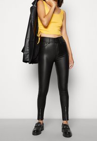 Černé skinny kalhoty z umělé kůže s vysokým pasem a zipovými detaily na kotnících, spojené s žlutým přiléhavým tílkem a černou koženou bundou.