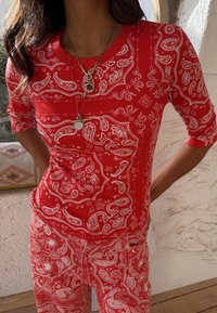 Γυναίκα ντυμένη με κόκκινη μπλούζα και παντελόνι με πασλεί μοτίβο, φορώντας στρωματωτά χρυσά κολιέ, στέκεται με τα χέρια στη μέση μέσα σε κλειστό χώρο.